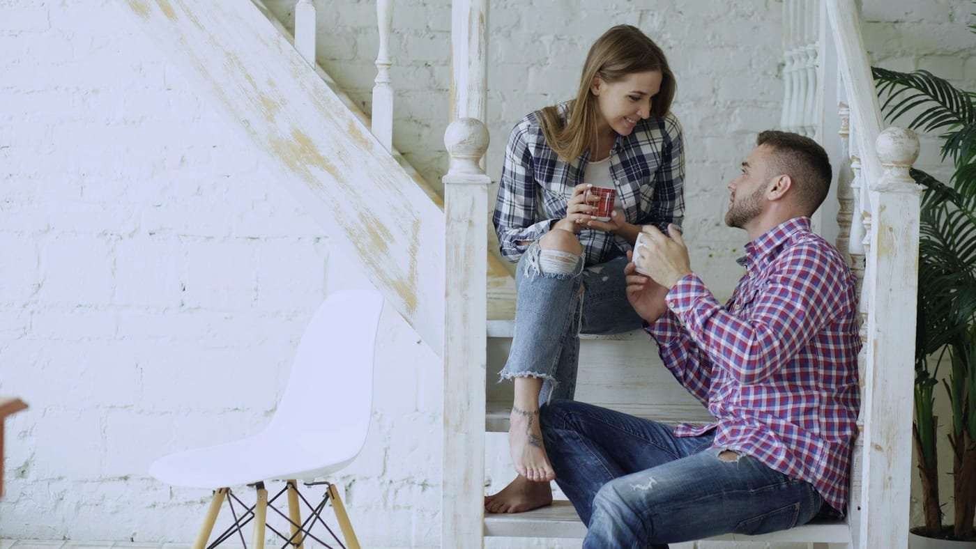 Casal conversando sentado na escada de casa, cada um com uma caneca nas mãos.